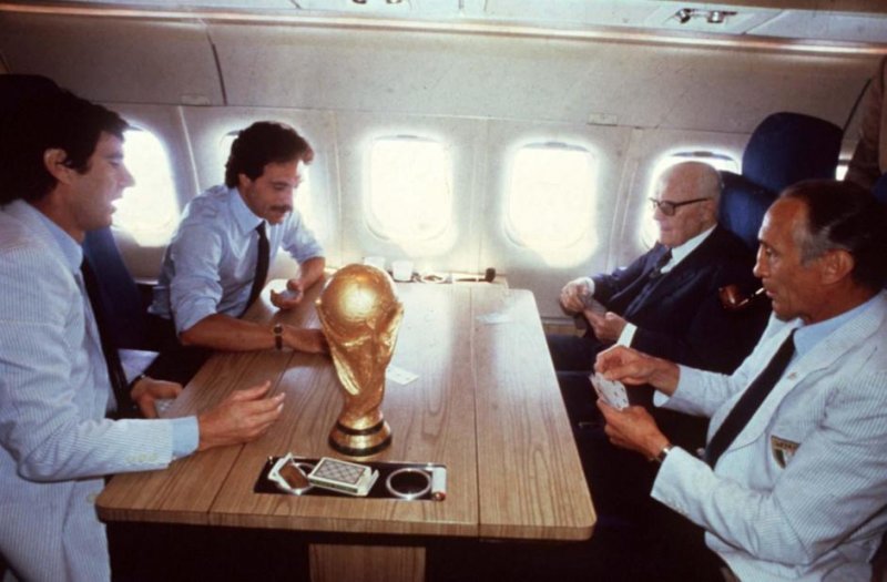Quattro personaggi illustri della storia italiana giocano a carte su un aereo, sul tavolo la coppa dei mondiali